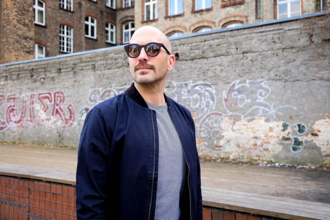Moody trägt vor einer Kreuzberger Hauswand eine GOBI Amsterdam Sonnenbrille zu T-Shirt und Blouson