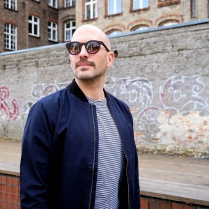 Moody trägt vor einer Kreuzberger Hauswand eine GOBI Amsterdam Sonnenbrille zu T-Shirt und Blouson